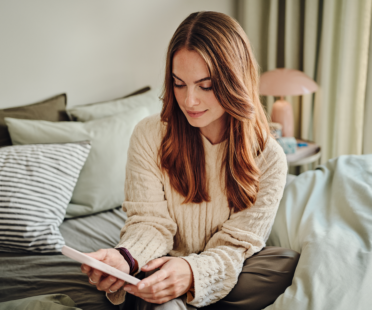 A woman sitting up in bed, smiling at the Natural Cycles app on her phone, wearing the NC Band